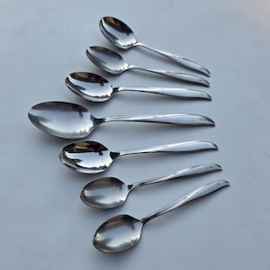 May include: A set of seven silver-toned metal spoons arranged in a vertical row. Each spoon features a smooth, polished bowl and a handle decorated with a small, etched star-like pattern near the end. The spoons vary slightly in size.