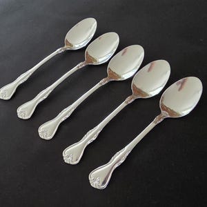 May include: Five silver spoons arranged diagonally on a dark surface. Each spoon has a long, slender handle with ornate detailing and a rounded bowl. The spoons reflect the light, creating a bright, shiny appearance. The spoons are likely for serving or eating.