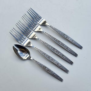 Peut inclure: Un ensemble de cinq fourchettes et une cuillère en acier inoxydable. Les couverts présentent un motif floral décoratif sur les manches. Les fourchettes ont quatre dents chacune. La cuillère a un bol arrondi.