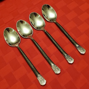 May include: Four silver spoons with hammered black handles and ornate, engraved designs on the ends. The spoons have a reflective, polished surface and are arranged diagonally on a red tablecloth.