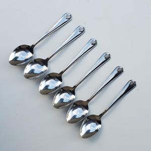 May include: Six shiny, silver spoons arranged diagonally on a white surface. Each spoon has a rounded bowl and a long handle with a decorative flourish at the top. The spoons reflect the surrounding environment.