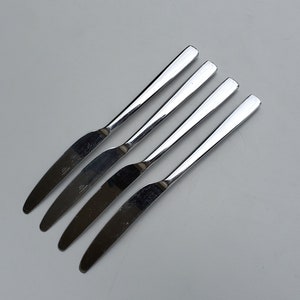 May include: Four silver dinner knives laid out in a row on a white surface.