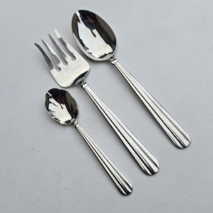 May include: A set of three polished silver serving utensils: a large spoon, a fork, and a smaller spoon. The handles have a ribbed design, and the utensils are arranged diagonally against a light gray background.