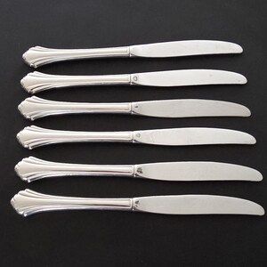 May include: Six silver butter knives with decorative handles are arranged on a black surface. The knives have a classic design with a rounded blade and a detailed handle. The blades are smooth and shiny.
