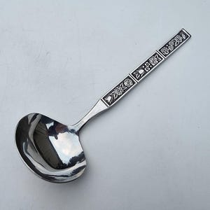 May include: A silver ladle with a floral design on the handle. The ladle has a rounded bowl and a long handle with engraved flowers and geometric shapes. The metal is shiny and reflects light.