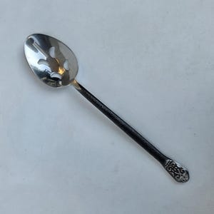 May include: A silver-toned slotted spoon featuring a polished, oval-shaped bowl with four cut-out openings. The handle is dark, textured, and matte, ending in a decorative, embossed silver-toned tip with an intricate, symmetrical scroll pattern.