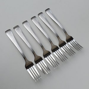 May include: Seven stainless steel forks arranged diagonally on a light gray surface. Each fork has a long, slender handle with a squared-off end and four tines. The forks are evenly spaced and reflect the surrounding light.