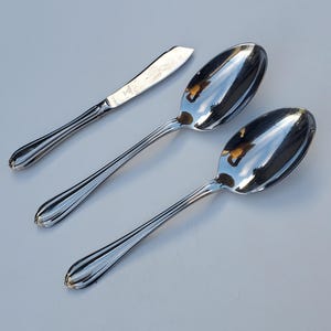 May include: A set of three polished silver utensils: a butter knife and two spoons. The knife has a rounded blade and a ridged handle. The spoons have oval bowls and matching ridged handles. The utensils are arranged diagonally on a light-colored surface.