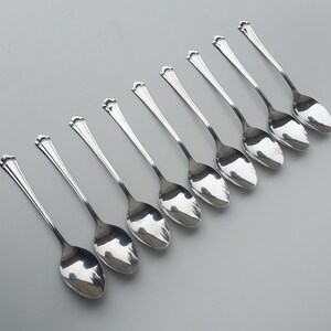 May include: A set of eight silver spoons arranged diagonally on a gray surface. Each spoon has a rounded bowl and a handle that flares out to a decorative, three-lobed design at the top. The spoons reflect the light, creating highlights.