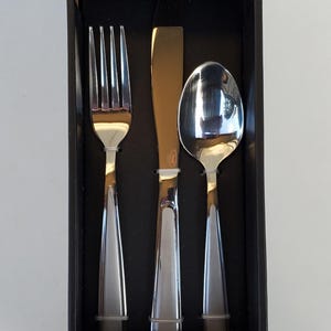 May include: A boxed set of Oneida Jordan silverware. The set includes a fork, knife, and spoon. The knife has a gold-colored blade. The box is black with the Oneida and Jordan brand names. The set is for 4 people and includes 12 pieces.