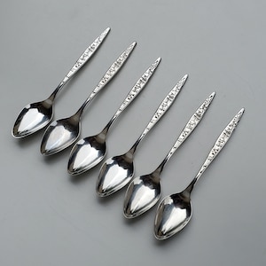 May include: Six silver spoons with an ornate design. The spoons are arranged in a row, with the handles pointing to the right.