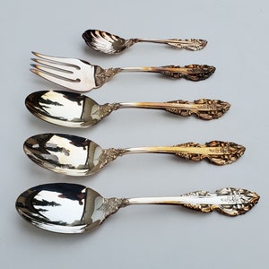 May include: A collection of ornate silver serving utensils, including a small spoon, a fork, and three spoons. The utensils have detailed floral and scrollwork designs on the handles and are arranged on a light gray surface.