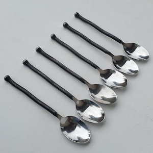 May include: Six spoons with a dark, textured handle and a shiny, oval-shaped silver bowl. The spoons are arranged diagonally across a light gray surface, showcasing their elegant design and craftsmanship.
