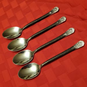 May include: Four silver spoons with hammered black handles and decorative ends. The spoons are arranged diagonally on a red checkered tablecloth. The spoons reflect light.
