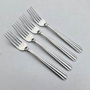 May include: Four stainless steel forks arranged diagonally across a light gray surface. Each fork has four tines and a handle with a decorative, ridged design. The forks are evenly spaced and appear to be new.