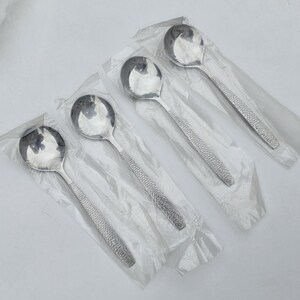 May include: Four silver-toned metal spoons, each individually wrapped in clear plastic packaging. The spoons feature a polished, reflective bowl and a textured handle with a repeating circular pattern along the length of the stem.