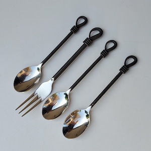 May include: Set of four handcrafted spoons and a fork. The spoons and fork have shiny silver bowls and dark metal handles with a looped design at the top. The utensils are arranged diagonally on a light surface.