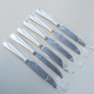 May include: Set of six silver dinner knives in individual plastic packaging. The knives have a beaded handle and a plain blade.