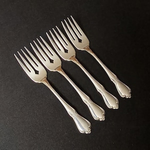 May include: Four silver forks with decorative handles and diamond-shaped cutouts on the tines. The forks are arranged diagonally against a dark background, showcasing their elegant design and polished finish.