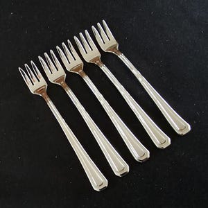 May include: Six silver forks arranged diagonally on a black surface. Each fork has four tines and a long, faceted handle. The forks are made of a shiny, reflective metal.
