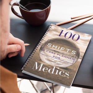 Peut inclure: Un agenda à spirales intitulé "100 Shifts A Care Planner For Medics" avec une couverture de scène de plage. Une tasse marron avec une cuillère et des crayons sont sur une surface sombre. Une personne tient un crayon.