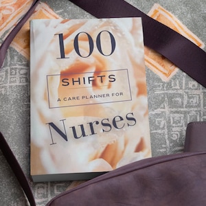 Op de afbeelding: Een wit boek met een bloemenontwerp op de omslag. De titel van het boek is "100 Shifts: A Care Planner for Nurses".