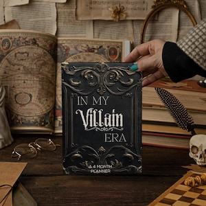 May include: A black and gold planner with the text "IN MY Villain ERA" is held by a person. The planner is surrounded by books, a bust, and a skull. The planner is a 4-month planner.