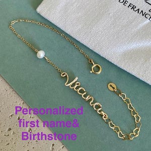 Puede incluir: Una pulsera dorada con el nombre "Vianca" en escritura cursiva, una perla y una cadena en forma de corazón. La pulsera se muestra sobre una superficie verde azulado, junto a una bolsa blanca con el texto negro "Beloy Bijoux DE FRANCE". El texto en la imagen dice: "Nombre personalizado y piedra de nacimiento".