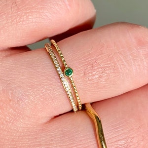 Peut inclure: Une bague en or avec une petite pierre précieuse vert émeraude sertie au centre. La bague est empilée avec deux autres bagues en or, l'une avec une rangée de petits diamants et l'autre avec une bande texturée.