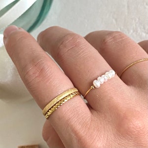 Peut inclure: Deux bagues en or sur un doigt. La bague du haut est une chaîne délicate avec cinq petites perles blanches. La bague du bas est une bande épaisse avec une surface texturée.