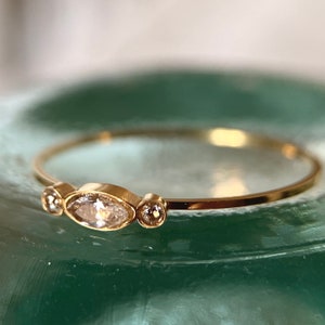 Peut inclure: Une délicate bague en or avec trois pierres précieuses transparentes. La pierre centrale est de forme ovale, et les deux plus petites pierres sont rondes. La bague est posée sur une surface verte.