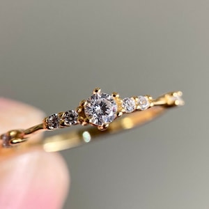 Op de afbeelding: Een delicate gouden ring met een enkele, ronde, heldere edelsteen omgeven door kleinere, heldere edelstenen. De ring heeft een delicaat, rankvormig ontwerp.
