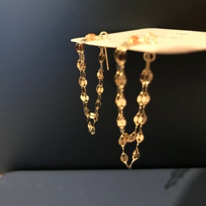 Peut inclure: Une paire de boucles d'oreilles en chaîne dorée avec de petits maillons ronds. Les boucles d'oreilles sont suspendues à une carte blanche.