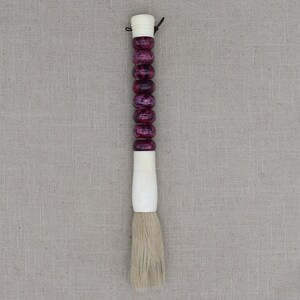 K&ouml;nnte beinhalten: Ein wei&szlig;er und lila Pinsel mit einem langen Griff aus Perlen. Der Pinsel besteht aus Naturborsten und wird f&uuml;r Kalligraphie verwendet.