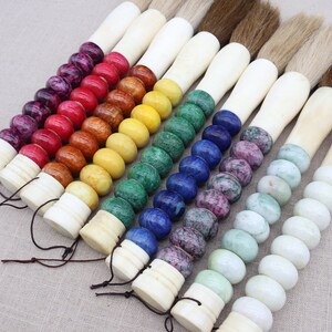 K&ouml;nnte beinhalten: Ein Set aus sieben Kalligraphie-Pinseln mit Knochengriffen, die mit bunten Edelsteinperlen verziert sind. Die Perlen sind in einem Regenbogenmuster angeordnet, beginnend mit Lila und endend mit Gr&uuml;n.