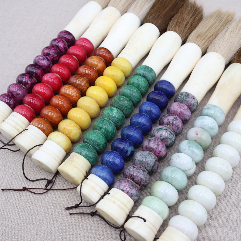 K&ouml;nnte beinhalten: Ein Set aus sieben Kalligraphie-Pinseln mit Knochengriffen, die mit bunten Edelsteinperlen verziert sind. Die Perlen sind in einem Regenbogenmuster angeordnet, beginnend mit Lila und endend mit Gr&uuml;n.