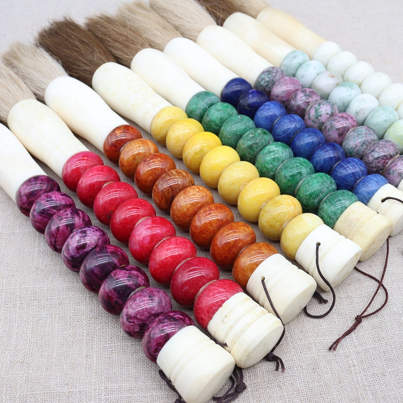 K&ouml;nnte beinhalten: Ein Set aus zehn Kalligraphie-Pinseln mit bunten, runden Perlen an den Griffen. Die Perlen sind in einem Regenbogenmuster angeordnet, beginnend mit Lila und endend mit Gr&uuml;n. Die Pinsel sind aus nat&uuml;rlichen Materialien gefertigt und haben ein traditionelles Design.