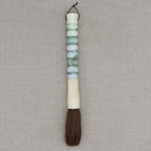 K&ouml;nnte beinhalten: Ein traditioneller chinesischer Kalligraphie-Pinsel mit einem langen, elfenbeinfarbenen Griff. Der Griff ist mit einer Reihe von jadegr&uuml;nen Perlen verziert. Der Pinsel hat eine lange, dunkelbraune Borste.