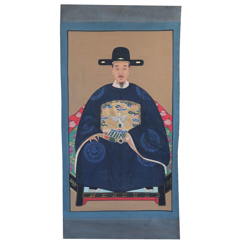 Pode incluir: Retrato de um homem em traje asi&aacute;tico tradicional. Ele usa uma t&uacute;nica azul escura com detalhes ornamentados, um chap&eacute;u preto e est&aacute; sentado em uma cadeira. O fundo &eacute; bege com uma borda azul.