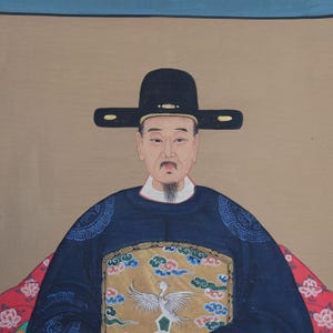 Pode incluir: Um retrato de um homem em trajes asi&aacute;ticos tradicionais. Ele usa um chap&eacute;u preto e uma t&uacute;nica azul escura com bordados dourados e azuis. O fundo &eacute; de cor bege neutra, com uma borda azul na parte superior.