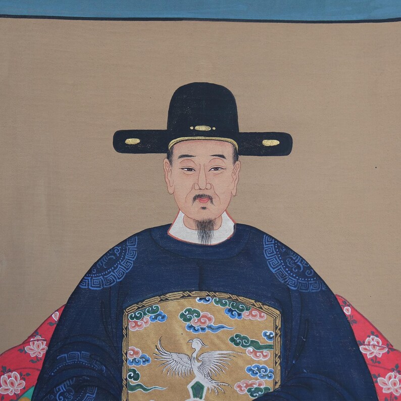 Pode incluir: Um retrato de um homem em trajes asi&aacute;ticos tradicionais. Ele usa um chap&eacute;u preto e uma t&uacute;nica azul escura com bordados dourados e azuis. O fundo &eacute; de cor bege neutra, com uma borda azul na parte superior.