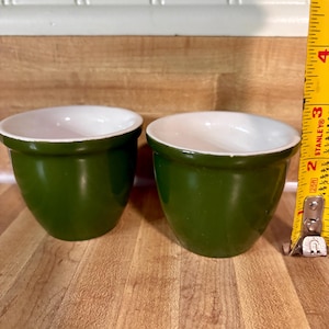 May include: Two small, round, dark green bowls with white interiors. A yellow Stanley measuring tape is positioned vertically next to the right bowl, indicating a height of approximately 2.5 inches for the bowls.