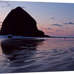 May include: A scenic landscape photograph of a beach at dusk. A large, dark rock formation dominates the left side, silhouetted against a sky of pink, orange, and blue hues. The wet sand reflects the colors of the sky.