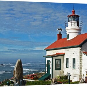 May include: A white stone lighthouse with a bright red roof and a lantern room sits on a rocky coastline overlooking the blue ocean under a partly cloudy sky. The structure features dark green window frames, a small red picnic table, and two chimneys.