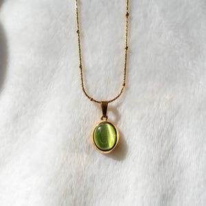 Op de afbeelding: Een goudkleurige ketting met een fijne schakelketting met kleine, gelijkmatig verdeelde kraaltjes en een ronde hanger. De hanger bevat een gladde, doorschijnende, lichtgroene cabochonsteen in een gepolijste goudkleurige metalen zetting.