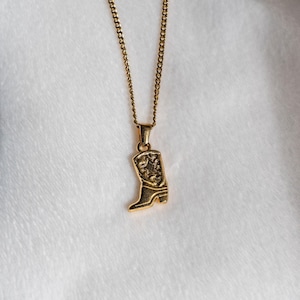 May include: A gold-toned metal necklace featuring a delicate chain and a pendant shaped like a cowboy boot. The boot pendant has a textured surface with a star detail on the shaft and a small heel, suspended from a simple jump ring bail.