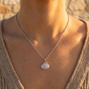 Può includere: Una collana d'argento con un ciondolo a forma di conchiglia. Il ciondolo è impreziosito da piccoli dettagli scintillanti. La collana è indossata su un maglione dai toni neutri, creando un look semplice ed elegante. La catena è delicata.