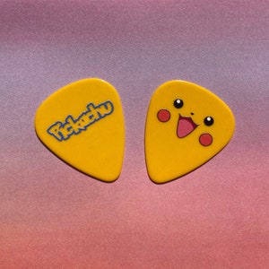 Könnte beinhalten: Zwei gelbe Gitarrenplektren mit einem Pikachu-Design. Ein Plektrum hat den Schriftzug "Pikachu" in blauen Buchstaben. Das andere Plektrum zeigt ein Cartoon-Bild von Pikachus Gesicht.