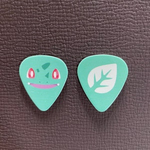 Könnte beinhalten: Zwei hellgrüne Gitarrenplektren. Eines zeigt ein Cartoon-Gesicht mit roten Augen und einem rosa Mund. Das andere hat ein weißes Blattdesign. Beide Plektren liegen auf einer braunen, strukturierten Oberfläche.