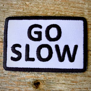 May include: White embroidered patch with black stitching. The patch has a black border and says "GO SLOW" in all caps.